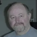 A middle-aged man with a beard and short hair is looking directly at the camera. About him, a dimly lit background features blurred objects, adding an air of mystery to his calm demeanor in a gray shirt.