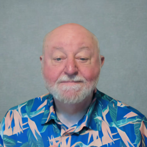 An older man with a white beard and bald head wearing a colorful, patterned shirt sits in front of a plain gray background. An older man with a white beard and bald head wearing a colorful, patterned shirt sits in front of a plain gray background.