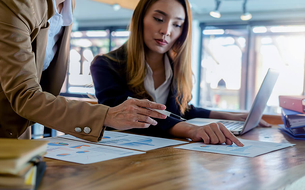 Two professionals review charts and graphs on paper at a desk, providing an honest review of legit online income opportunities; one points to a document while the other works on a laptop.