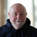 An older man with a white beard and mustache wears a navy hoodie and looks at the camera, with a blurred indoor background.